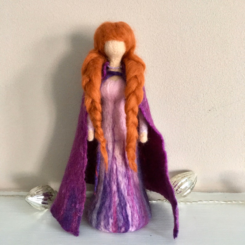 May include: A felt doll with long, braided red hair wearing a purple and pink dress and a purple cape.