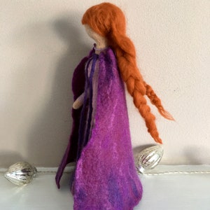 May include: A felt doll with long, braided red hair wearing a purple cape. The doll is standing on a white surface.