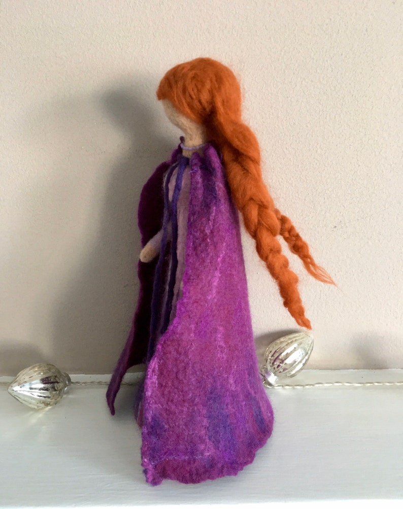 May include: A felt doll with long, braided red hair wearing a purple cape. The doll is standing on a white surface.