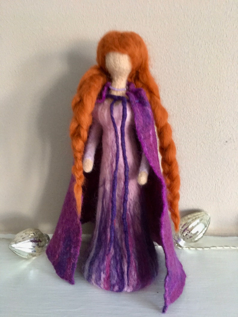 May include: A felt doll with long, braided red hair wearing a purple and pink dress and a purple cape.