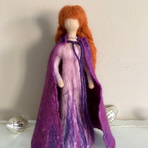 May include: A felt doll with long, red hair wearing a purple dress and a matching cape. The doll has a simple, white face.