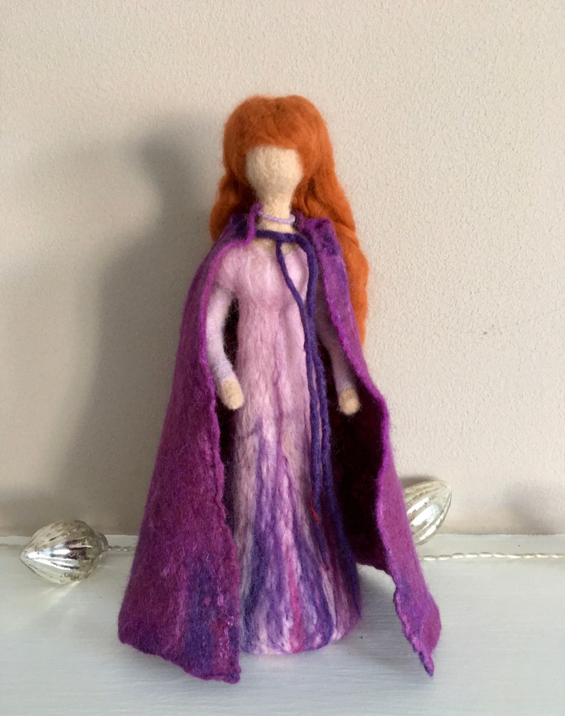 May include: A felt doll with long, red hair wearing a purple dress and a matching cape. The doll has a simple, white face.