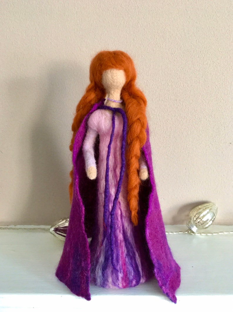 May include: A felt doll with long, braided red hair wearing a purple dress and a matching cape.
