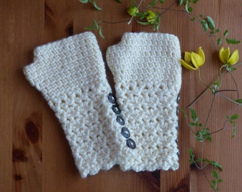 May include: A pair of ivory-colored, crocheted fingerless gloves. The gloves have a scalloped edge and are adorned with four dark, decorative buttons. The gloves are displayed on a wooden surface, with yellow flowers and green vines in the background.