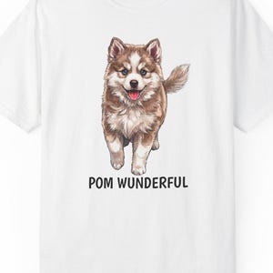 Pode incluir: T-shirt branco com uma ilustração em aquarela de um cachorrinho marrom e branco. O texto "POM WUNDERFUL" está impresso em preto abaixo. O cachorrinho está com a língua de fora. A camisa tem gola redonda.