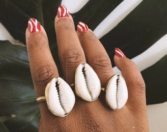 Puede incluir: Tres anillos dorados, cada uno con una concha blanca, se muestran en una mano con arte de uñas rojo y blanco. Los anillos se colocan sobre un fondo de una gran hoja verde y una superficie blanca.