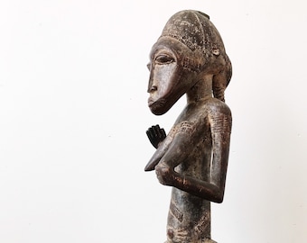 May include: A dark wood sculpture of a seated figure with a headdress. The figure has a detailed face and body with intricate carvings. The sculpture is sitting on a wooden base.