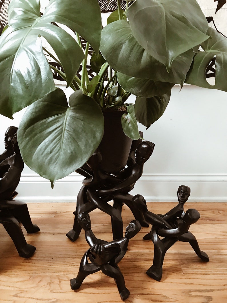 May include: A black wooden plant stand with a group of figures holding hands, supporting a potted plant with large green leaves.