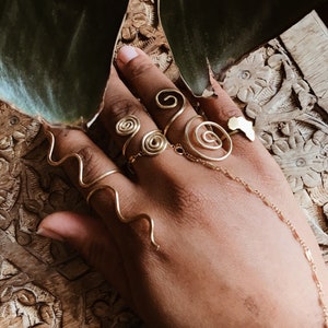 Pode incluir: Uma m&atilde;o adornada com an&eacute;is de arame dourado. Os an&eacute;is apresentam desenhos em espiral e ondulados, com um anel em forma de continente africano. Uma delicada corrente conecta os an&eacute;is.