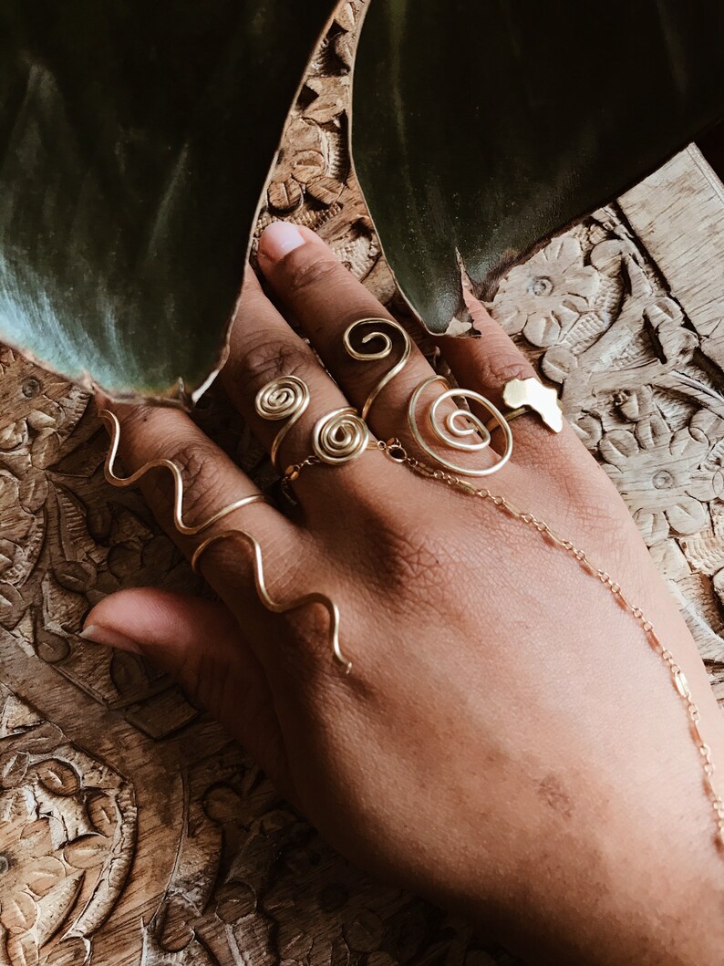 Pode incluir: Uma m&atilde;o adornada com an&eacute;is de arame dourado. Os an&eacute;is apresentam desenhos em espiral e ondulados, com um anel em forma de continente africano. Uma delicada corrente conecta os an&eacute;is.