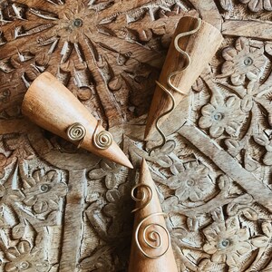 Pode incluir: Tr&ecirc;s suportes de anel de madeira com an&eacute;is de arame dourado. Os an&eacute;is apresentam desenhos em espiral e ondulados. Os suportes s&atilde;o exibidos em uma superf&iacute;cie de madeira entalhada.