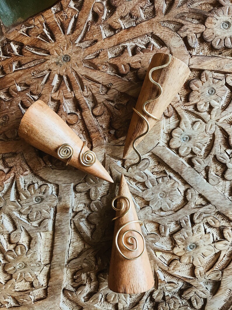 Pode incluir: Tr&ecirc;s suportes de anel de madeira com an&eacute;is de arame dourado. Os an&eacute;is apresentam desenhos em espiral e ondulados. Os suportes s&atilde;o exibidos em uma superf&iacute;cie de madeira entalhada.