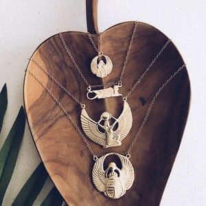 Puede incluir: Cuatro collares de oro con diferentes colgantes de tem&aacute;tica egipcia, incluyendo un escarabajo, una diosa alada y un ankh alado. Los collares se muestran sobre una superficie de madera.
