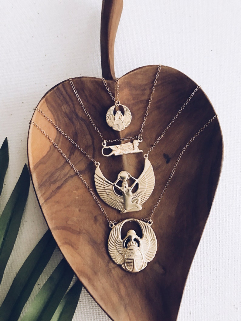 Puede incluir: Cuatro collares de oro con diferentes colgantes de tem&aacute;tica egipcia, incluyendo un escarabajo, una diosa alada y un ankh alado. Los collares se muestran sobre una superficie de madera.