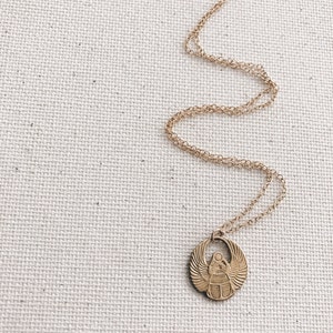 Puede incluir: Un collar de cadena dorada con un colgante de escarabajo. El colgante es redondo y presenta un dise&ntilde;o detallado de un escarabajo con alas.