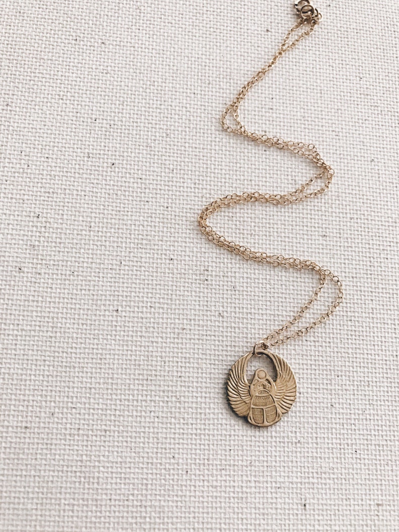 Puede incluir: Un collar de cadena dorada con un colgante de escarabajo. El colgante es redondo y presenta un dise&ntilde;o detallado de un escarabajo con alas.