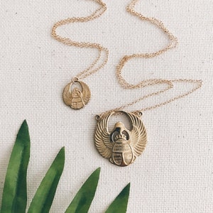 Puede incluir: Dos collares de oro con colgantes de escarabajo. El colgante m&aacute;s grande es un dise&ntilde;o detallado con alas y un cuerpo. El colgante m&aacute;s peque&ntilde;o es un dise&ntilde;o m&aacute;s simple. Los collares est&aacute;n sobre un fondo blanco con una hoja verde.