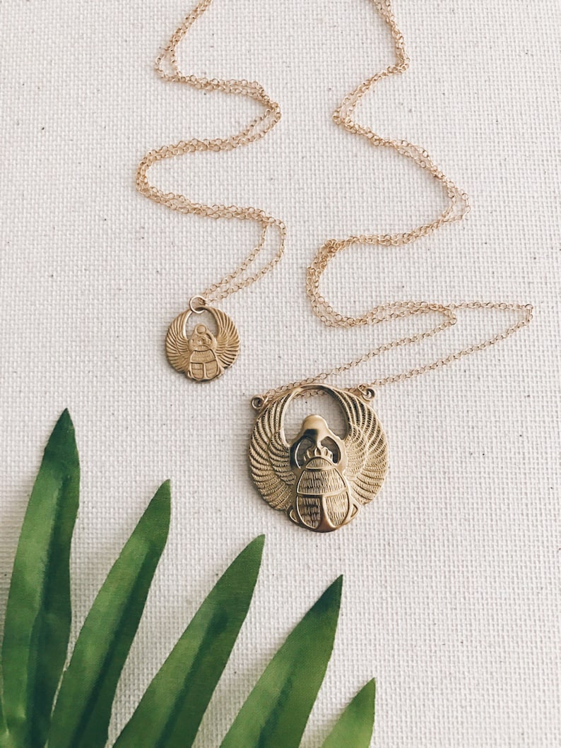 Puede incluir: Dos collares de oro con colgantes de escarabajo. El colgante m&aacute;s grande es un dise&ntilde;o detallado con alas y un cuerpo. El colgante m&aacute;s peque&ntilde;o es un dise&ntilde;o m&aacute;s simple. Los collares est&aacute;n sobre un fondo blanco con una hoja verde.