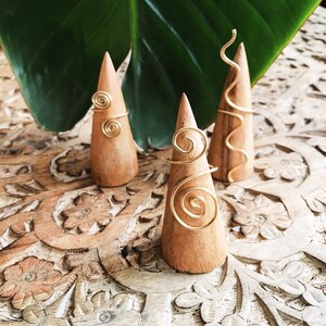 Pode incluir: Tr&ecirc;s suportes de anel de madeira com an&eacute;is de arame dourado. Os an&eacute;is apresentam designs em espiral e curvos. Os suportes s&atilde;o exibidos em uma superf&iacute;cie de madeira esculpida, com uma grande folha verde ao fundo.