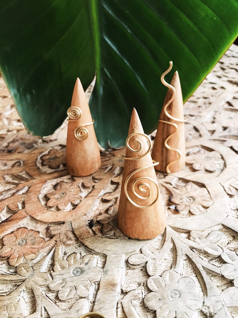 Pode incluir: Tr&ecirc;s suportes de anel de madeira com an&eacute;is de arame dourado. Os an&eacute;is apresentam designs em espiral e curvos. Os suportes s&atilde;o exibidos em uma superf&iacute;cie de madeira esculpida, com uma grande folha verde ao fundo.