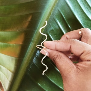 Pode incluir: Um par de brincos dourados em forma de onda. Os brincos s&atilde;o segurados contra uma grande folha verde. Os brincos t&ecirc;m um design simples e minimalista, com um acabamento liso e polido. S&atilde;o leves e delicados.