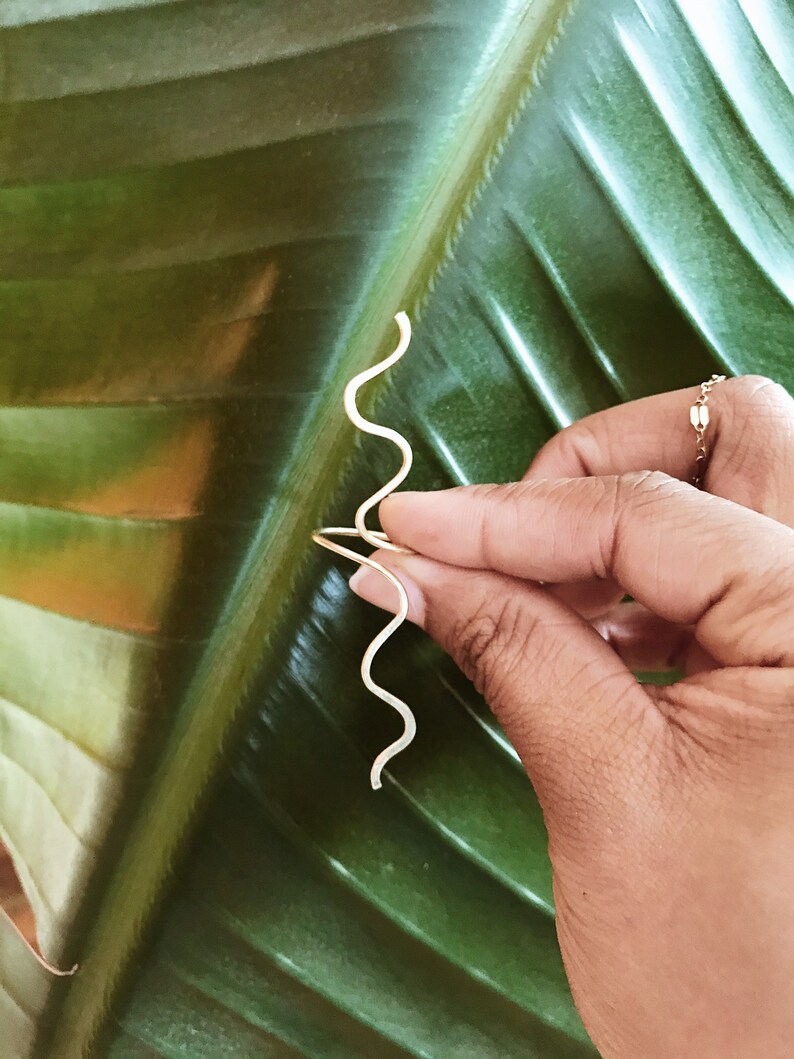 Pode incluir: Um par de brincos dourados em forma de onda. Os brincos s&atilde;o segurados contra uma grande folha verde. Os brincos t&ecirc;m um design simples e minimalista, com um acabamento liso e polido. S&atilde;o leves e delicados.