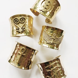 May include: Five gold-toned metal bracelets with different embossed symbols. The symbols include an Ankh, an Eye of Horus, a heart with a swirl design, a winged figure, and a symbol that looks like a stylized letter G.