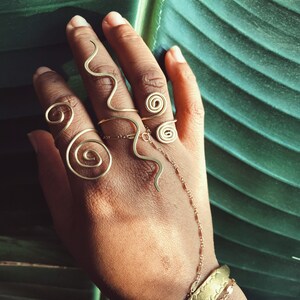 Pode incluir: Uma m&atilde;o adornada com v&aacute;rios an&eacute;is e pulseiras douradas. Os an&eacute;is apresentam desenhos em espiral e ondulados, ligados por uma delicada corrente. As pulseiras incluem uma pulseira larga e uma corrente fina. O fundo &eacute; uma grande folha verde.