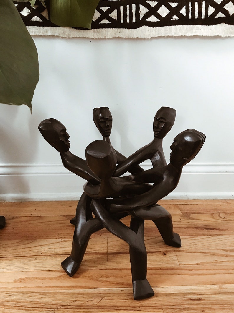May include: A wooden sculpture of five figures intertwined, forming a circular shape. The figures are carved in a dark brown color and have simple facial features. The sculpture is a unique and decorative piece of art.