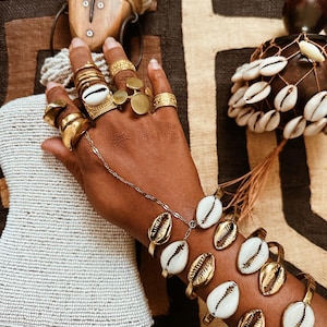 Könnte beinhalten: Eine Hand mit mehreren goldenen Ringen, darunter ein Ring mit einer Kaurimuschel. Eine feine Kette verbindet sich mit einem Armband mit Kaurimuscheln. Mehrere goldene Armbänder mit Kaurimuscheln sind ebenfalls zu sehen. Der Schmuck ist vor einem gemusterten Hintergrund platziert.