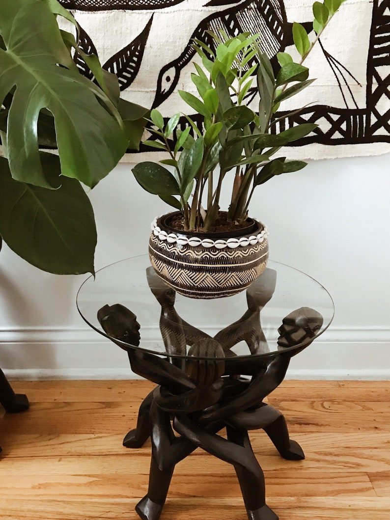 May include: A black wooden table with four legs carved in the shape of human figures. The table has a glass top and a potted plant with green leaves in a brown and white patterned pot.