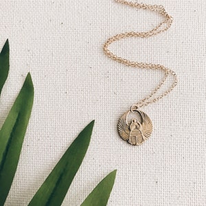 Puede incluir: Collar de cadena dorada con un colgante de escarabajo. El colgante es de dise&ntilde;o circular con el escarabajo en el centro y alas a cada lado. El collar est&aacute; sobre un fondo de tela blanca con hojas verdes en la esquina.