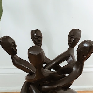 May include: A wooden sculpture of four figures intertwined, each with a carved face. The figures are dark brown and appear to be in a state of struggle or embrace.
