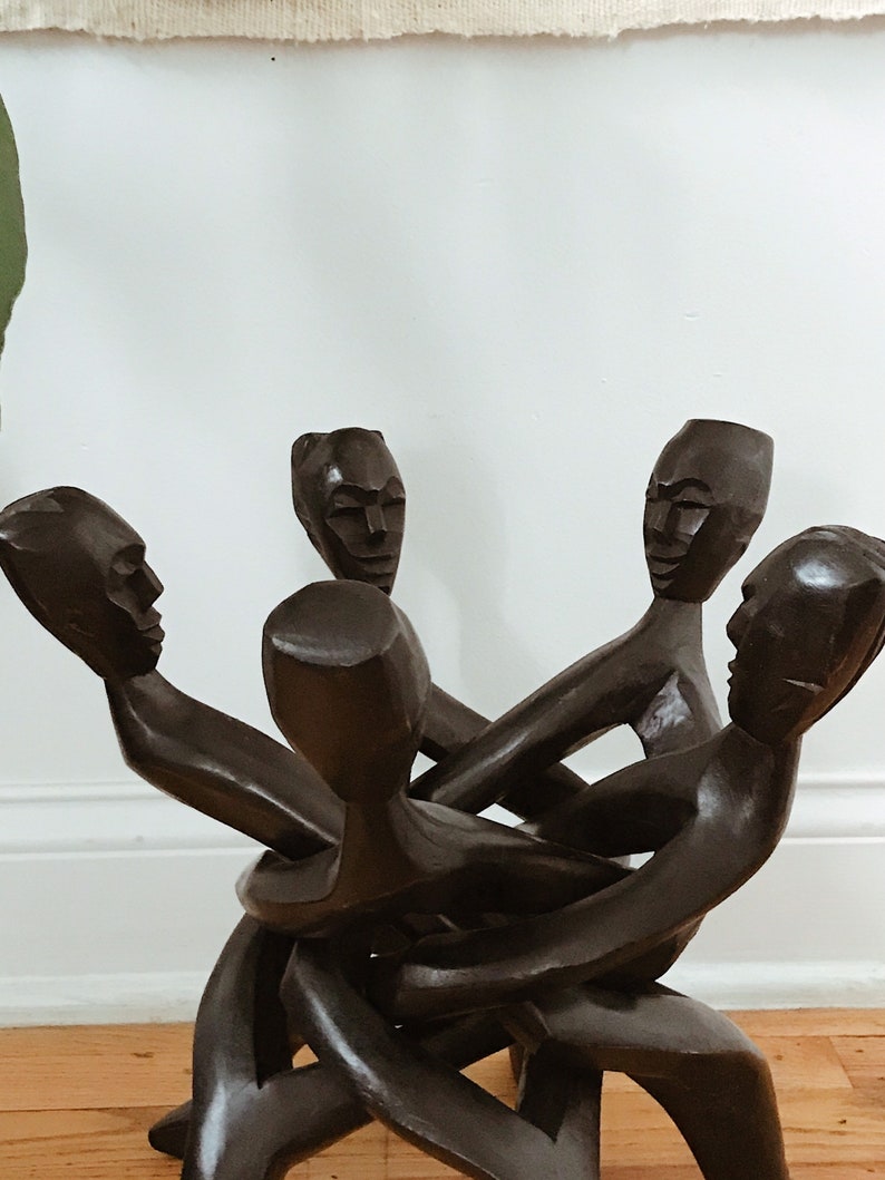 May include: A wooden sculpture of four figures intertwined, each with a carved face. The figures are dark brown and appear to be in a state of struggle or embrace.