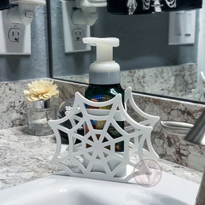 May include: White plastic spiderweb soap dispenser holder with a white bottle of soap inside. The holder is shaped like a spiderweb and has a small white skull on the top. The soap bottle is labeled "Bath & Body Works".