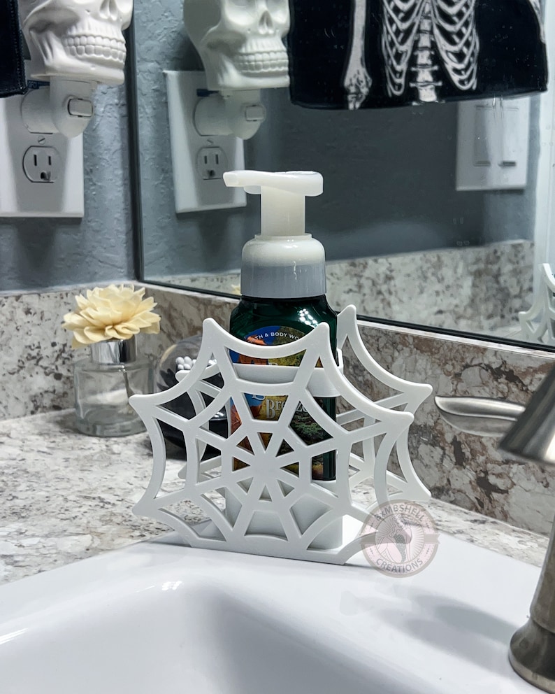 May include: White plastic spiderweb soap dispenser holder with a white bottle of soap inside. The holder is shaped like a spiderweb and has a small white skull on the top. The soap bottle is labeled "Bath & Body Works".