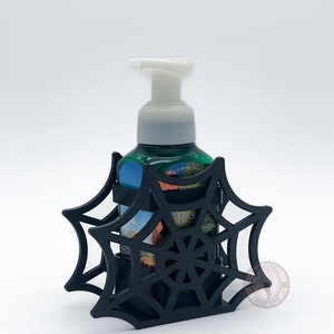May include: Black spiderweb-shaped soap dispenser holder with a green and white bottle of hand soap inside. The holder is made of plastic and has a black and white spiderweb design. The soap bottle is labeled "Bath & Body Works" and "Tree".