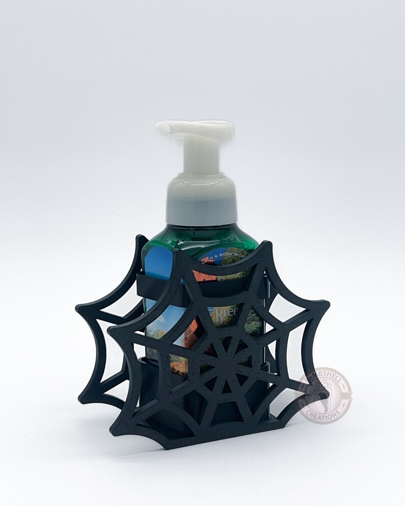 May include: Black spiderweb-shaped soap dispenser holder with a green and white bottle of hand soap inside. The holder is made of plastic and has a black and white spiderweb design. The soap bottle is labeled "Bath & Body Works" and "Tree".