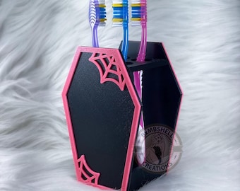 Op de afbeelding: Een zwarte en roze, kistvormige tandenborstelhouder met een spinnenwebontwerp. De houder heeft vier vakken voor tandenborstels. De tekst "XMBSHELL CREATIONS" staat aan de zijkant van de houder.