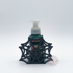 May include: Black spiderweb-shaped soap dispenser holder with a green and white bottle of foaming hand soap inside.