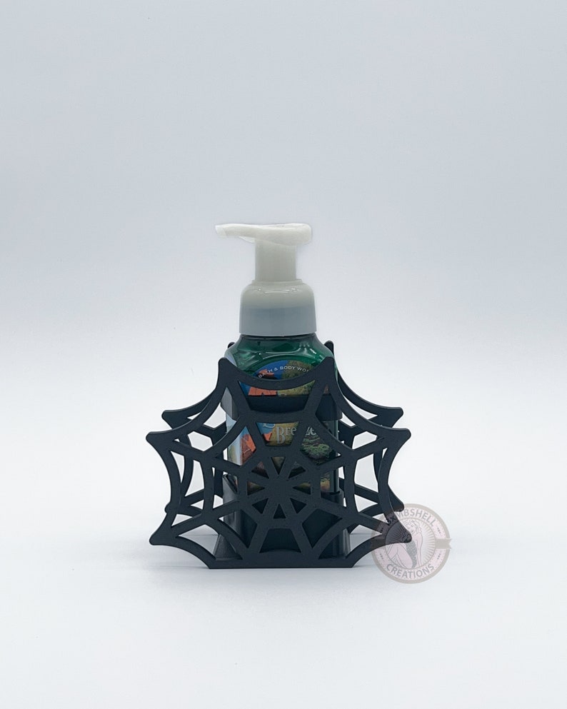 May include: Black spiderweb-shaped soap dispenser holder with a green and white bottle of foaming hand soap inside.