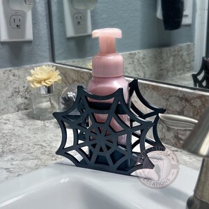 May include: Black spiderweb-shaped soap dispenser holder with a pink soap dispenser bottle inside. The holder is sitting on a white countertop in a bathroom.