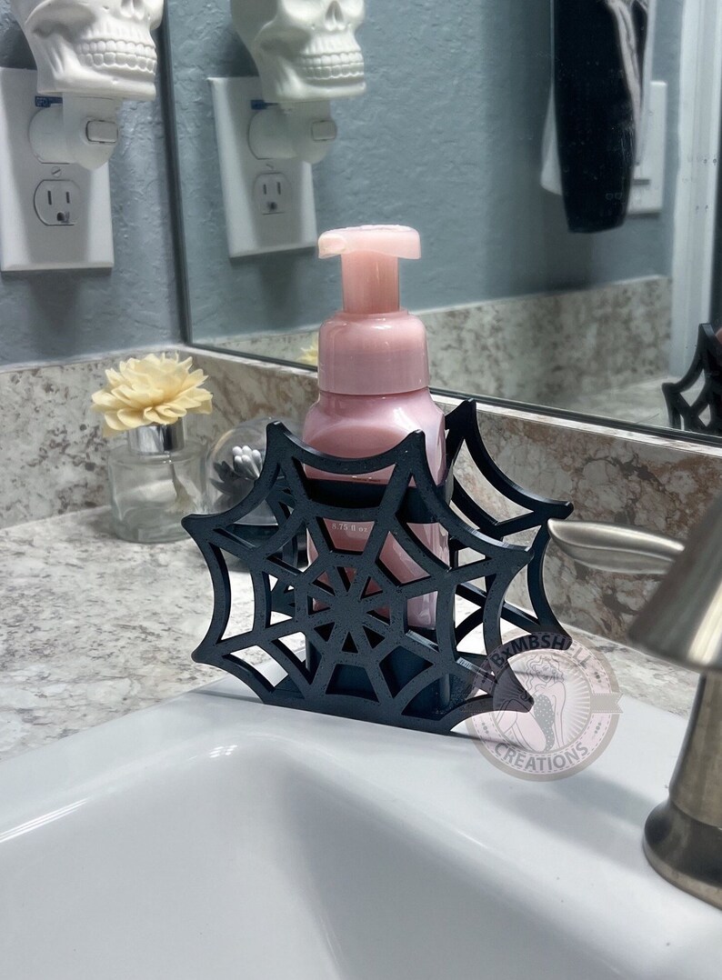 May include: Black spiderweb-shaped soap dispenser holder with a pink soap dispenser bottle inside. The holder is sitting on a white countertop in a bathroom.