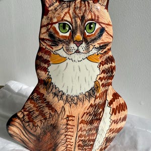May include: A ceramic figurine of a brown tabby cat with green eyes. The cat is sitting with its paws tucked underneath it. The figurine is hand-painted with details of the cat's fur and whiskers.