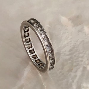 May include: A silver band ring with a row of clear square-shaped stones set around the entire band.