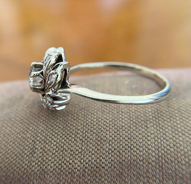 Puede incluir: Un anillo de plata con un dise&ntilde;o floral y piedras transparentes. El anillo est&aacute; sobre un fondo de tela marr&oacute;n.