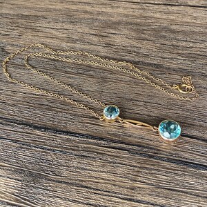 Op de afbeelding: Een gouden ketting met twee blauwe edelsteen hangers. De grotere hanger bevindt zich aan de rechterkant van de ketting en de kleinere hanger aan de linkerkant van de ketting.