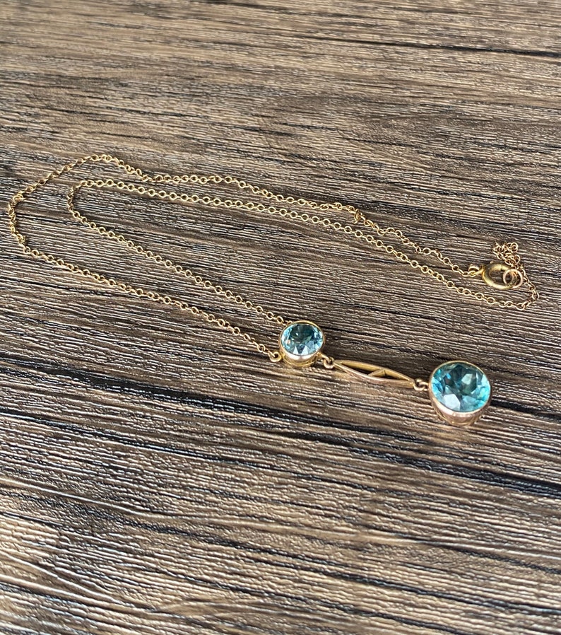 Op de afbeelding: Een gouden ketting met twee blauwe edelsteen hangers. De grotere hanger bevindt zich aan de rechterkant van de ketting en de kleinere hanger aan de linkerkant van de ketting.