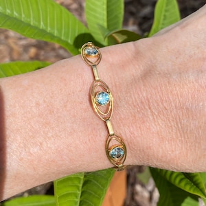 Op de afbeelding: Een gouden armband met drie ovale schakels, elk met een lichtblauwe edelsteen in het midden. De armband zit om een pols.