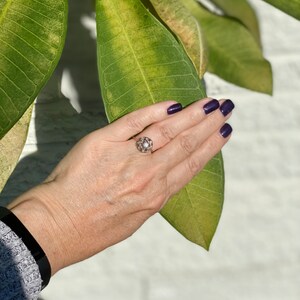 Puede incluir: Un anillo de oro con una piedra grande, redonda y transparente rodeada de piedras m&aacute;s peque&ntilde;as. El anillo est&aacute; en el dedo de una persona.